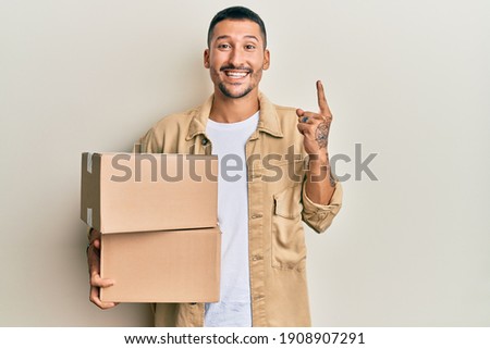 Handsome man with tattoos holding delivery package smiling with an idea or question pointing finger with happy face, number one 