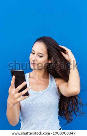 Portrait of a beautiful young woman smiling and using a smartphone with a blue wall in the background
