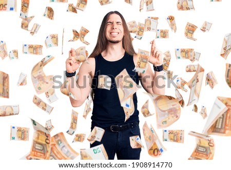 Young adult man with long hair wearing goth style with black clothes gesturing finger crossed smiling with hope and eyes closed. luck and superstitious concept.