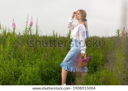 Beautiful blonde girl in a blue skirt and white shirt stands on among the green flowering meadows holding a wicker basket with flowers. High quality photo