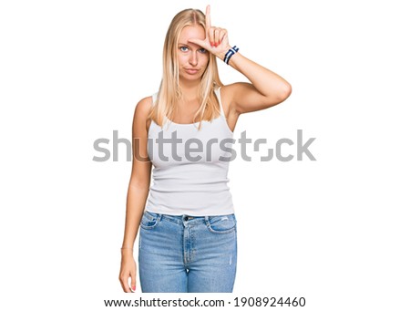 Young blonde girl wearing casual style with sleeveless shirt making fun of people with fingers on forehead doing loser gesture mocking and insulting. 