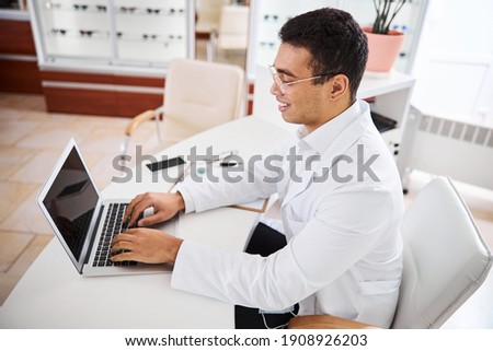 Smiling contented bespectacled eye doctor in a lab coat sitting before the laptop in his office Royalty-Free Stock Photo #1908926203