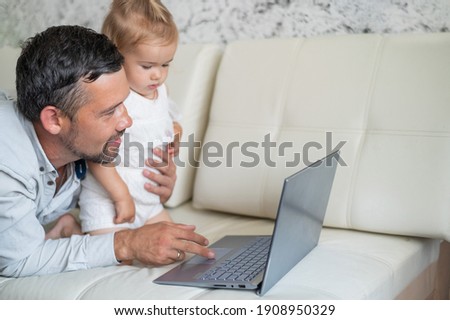 Little girl and caring father are watching cartoons on a laptop while lying on the couch at home. A man shows his little daughter photographs on a computer. Beloved daddy.