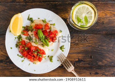 Halibut fish fillet baked with tomatoes. White plate, lemon, green basil, top view, close-up
