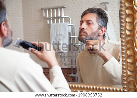 Trimming your beard with a trimmer. Nice gentleman in a beige T-shirt cuts his gray beard on his own using a typewriter in front of a large mirror in the bathroom

