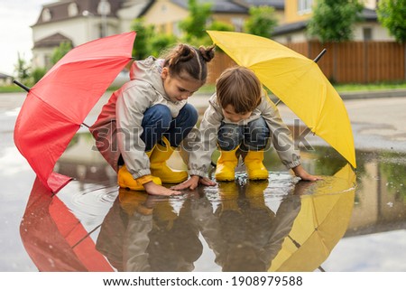Happy children play in a puddle after the rain. Brother and sister hid under umbrellas and touched water. Kids having fun on the street
