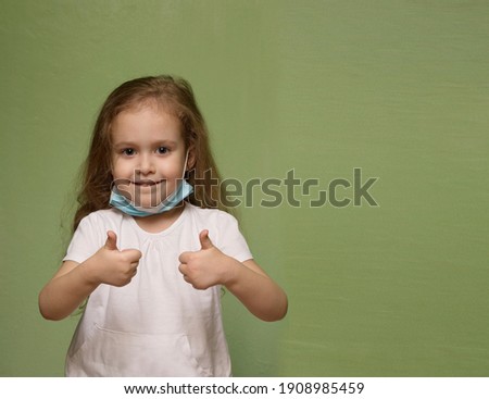 
A girl with a deflated medical mask shows thumbs up on a green background. Covid 19. Isolated photo of a child.