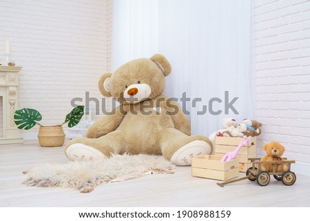 huge teddy bear sits in a bright room with toys in natural wicker basket with teddy bears, wooden cart and monstera leaves. Modern interior with light brick walls and wooden parquet floors. Copy space