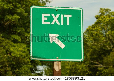 A simple green square shaped exit sign by a road. It has white full capital text with a tilted arrow showing the direction. There are trees in the background. A versatile image for getting out concept