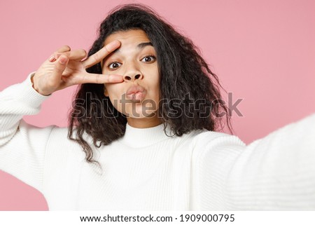 Close up young cute cheerful african american woman 20s in white knitted sweater do selfie shot on mobile phone show victory v-sign gesture duckface isolated on pastel pink background studio portrait.