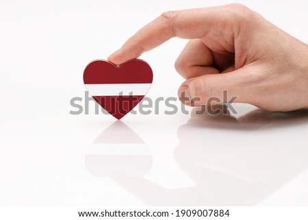 Love and respect Latvia. A man's hand holds a heart in the shape of the Latvian flag on a white glass surface. The concept of Latvian patriotism and pride.