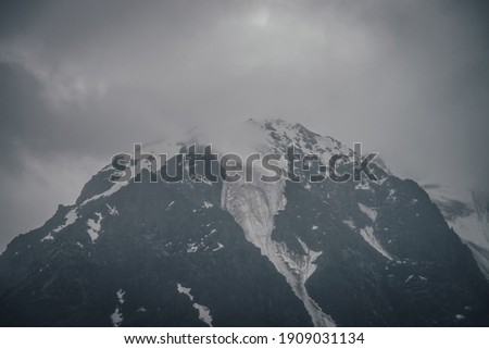 Dark atmospheric mountain landscape with glacier on black rocks in lead gray cloudy sky. Snowy mountains in low clouds in rainy weather. Gloomy landscape with black rocky mountains with snow in fog.