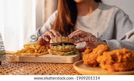 Closeup image of a woman holding and eating hamburger and french fries with fried chicken on the table at home Royalty-Free Stock Photo #1909201114
