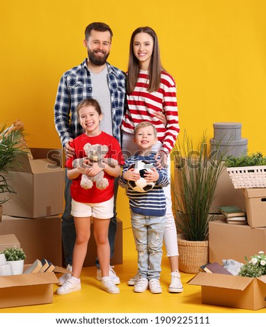 Full body of happy young parents with children looking at camera while standing together near unpacked boxes with belongings after moving in new house