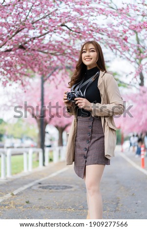 young woman traveler looking cherry blossoms or sakura flower blooming and holding camera to take a photo in the park