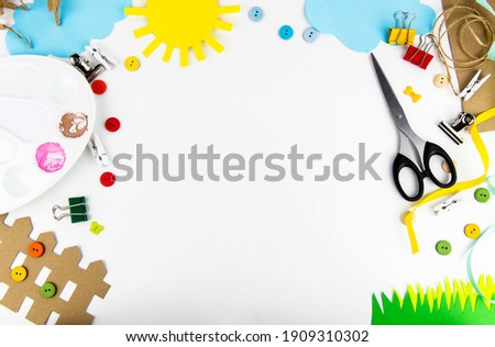 The background is a view from above on a white table with children's creativity. On the table of colored paper the sun, clouds, grass, fence, palette, scissors, colored buttons
