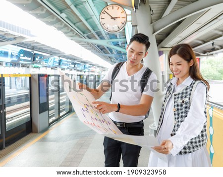 Portrait of cute smiling young adult Asian couple tourists standing and holding paper metro map together with man pointing to the map to show destination station with Skytrain station background