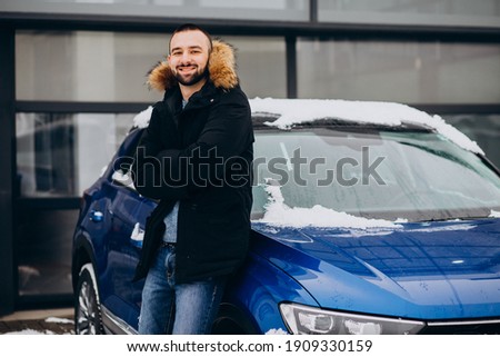 Handsome man in warm jacket standing by car covered with snow