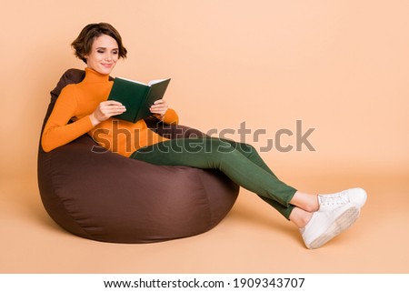 Full length photo of charming young girl orange turtleneck sitting bean bag reading book isolated beige color background