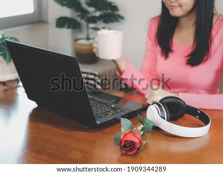 Cropped photo of woman holding cup of coffee, sitting at table with computer laptop, headphones and red rose. Selective focus on rose. woman working at computer on Valentine's day.
