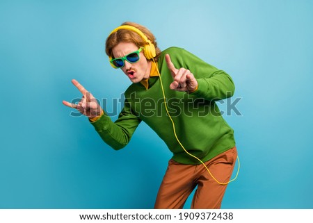 Photo of young funky funny cool brutal man in headphones showing rock n roll sign isolated on blue color background