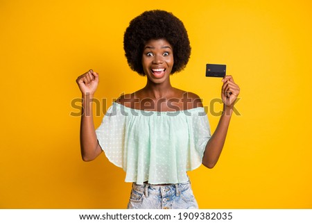 Photo portrait of excited girl celebrating with raised fist holding credit card in one hand isolated on vivid yellow colored background