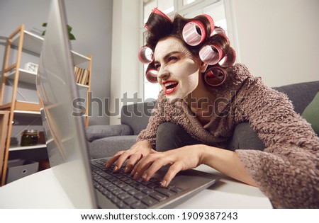 Mad woman in face mask and hair rollers sitting looking at laptop computer screen, writing insulting hater comment, spreading fake rumour. Jealous wife reading husband's direct message on social media Royalty-Free Stock Photo #1909387243