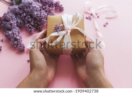 Happy mothers day and valentine's day concept. Hands holding gift box with ribbon and lilac flowers on pink paper. Purple lilac flowers bouquet with craft present box. Giving gift