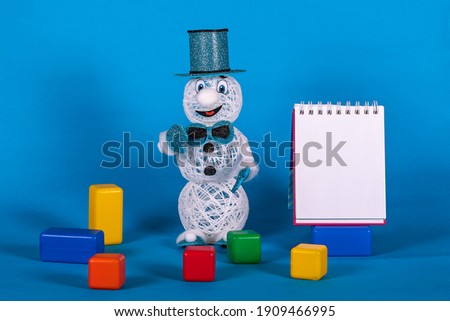 New Year's adventures, a snowman in a blue cilendra stands near a clean tablet for notes, colorful cubes lie around 