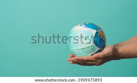 Two Hand is holding earth globe and face mask on green or Tiffany Blue background.covid-19 concept.