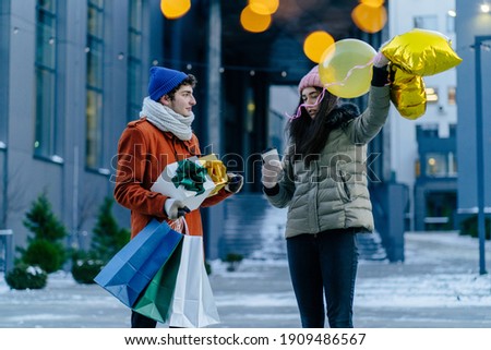 Young couple of male and female students in warm colorful outerwear holding gold balloons, gift boxes and paper bags outdoor in winter frozy day.