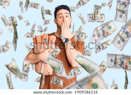 Young hispanic man holding paper bag with bread covering mouth with hand, shocked and afraid for mistake. surprised expression