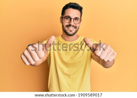 Young hispanic man wearing casual clothes and glasses approving doing positive gesture with hand, thumbs up smiling and happy for success. winner gesture.  Royalty-Free Stock Photo #1909589017