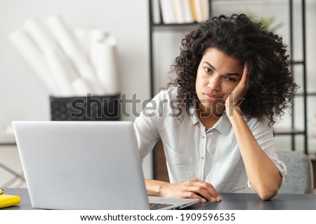 Young African American woman with afro hairstyle looks annoyed and stressed, sitting at the desk, using a laptop, thinking and looking at the camera, feeling tired and bored with depression problems