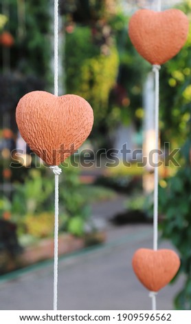 Closeup of brown heart-shape handmade earthenware hanging by white rope  with garden background in sunny day. Symbol of love and valentine day. Vertical view.
