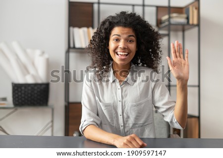 Young and energetic African-American female fresh graduate is ready for an online interview on a video call, sitting at the desk and looking and waving at the camera, saying hello. Job hunting concept