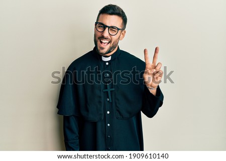 Young hispanic man wearing priest uniform standing over white background smiling with happy face winking at the camera doing victory sign. number two. 