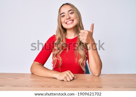 Young beautiful blonde woman wearing casual clothes sitting on the table smiling happy and positive, thumb up doing excellent and approval sign 