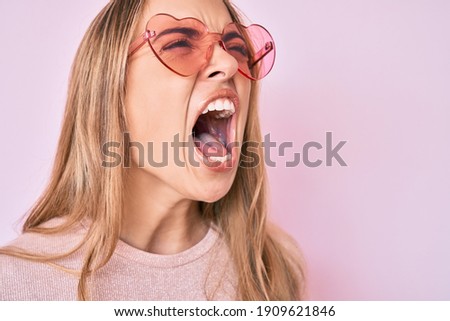 Young beautiful blonde woman wearing heart shaped sunglasses angry and mad screaming frustrated and furious, shouting with anger. rage and aggressive concept.  Royalty-Free Stock Photo #1909621846