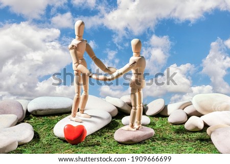 Two mannequins hold hands against a blue sky with white clouds, smooth cobblestones and a heart symbol in the foreground on green grass