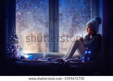 A little girl in a blue hat sits on the windowsill and looks at the snow. Next to her is a Christmas tree, a garland is burning, there is a candle