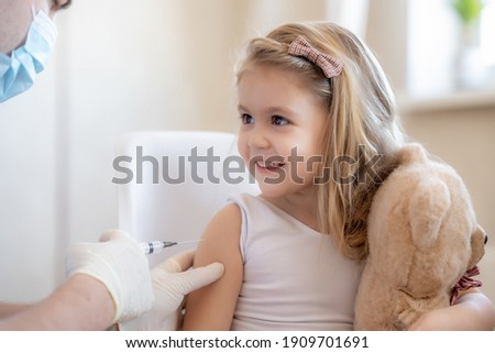 Happy little cute blonde girl holding a toy and getting a flu shot not afraid of the syringe needle. Doctor injecting brave child with Covid-19 vaccine at clinic
Immunization for children concept. 