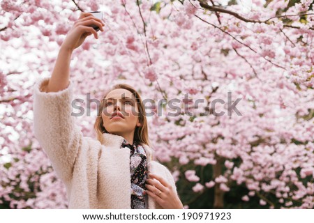 Beautiful young lady photographing nature with her mobile phone. Pretty young woman at spring blossom park taking photographs with her cell phone.