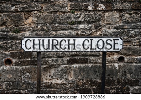 Traditional English Church Street postal address sign black text on white metal plate standing on foot path of road with stone church wall behind giving place name
