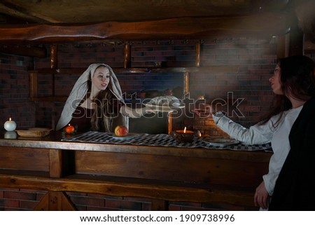 Women in historical costumes. The work of two maids in a tavern. A scene from medieval life. Historical reconstruction of a restaurant lunch. The girl serves bread to the maid. Renaissance painting. Royalty-Free Stock Photo #1909738996