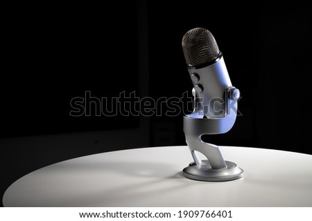 Silver grey USB condenser microphone with adjustable tabletop stand sitting on a white table in a dark contrast room with a neon light cyan highlight shine color pop with copy space