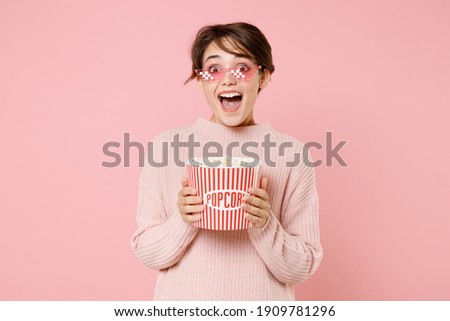 Excited young brunette woman wearing knitted casual sweater 8 bit glasses isolated on pastel pink background. People emotions in cinema, lifestyle concept. Watching movie film, hold bucket of popcorn