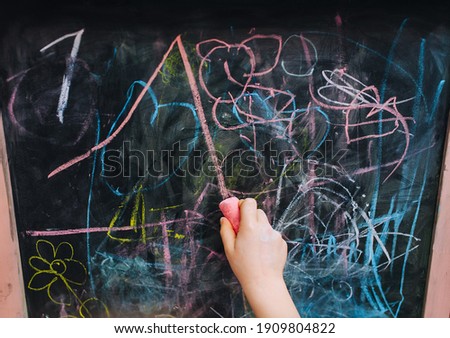 Preschool girl child draws, writes with the right hand with pink chalk on a black wooden board, easel a picture, scribbles and a straight line. Drawing lesson at school.
