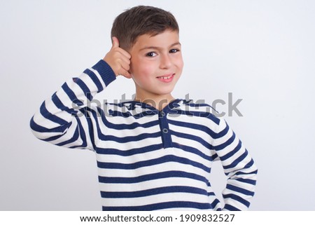little cute Caucasian boy kid wearing stripped t-shirt against white wall imitates telephone conversation, makes phone call gesture with hands, has confident expression. Call me!