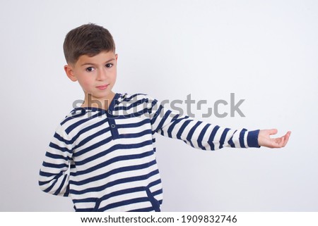 Portrait of little cute Caucasian boy kid wearing stripped t-shirt against white wall  with arm out in a welcoming gesture.